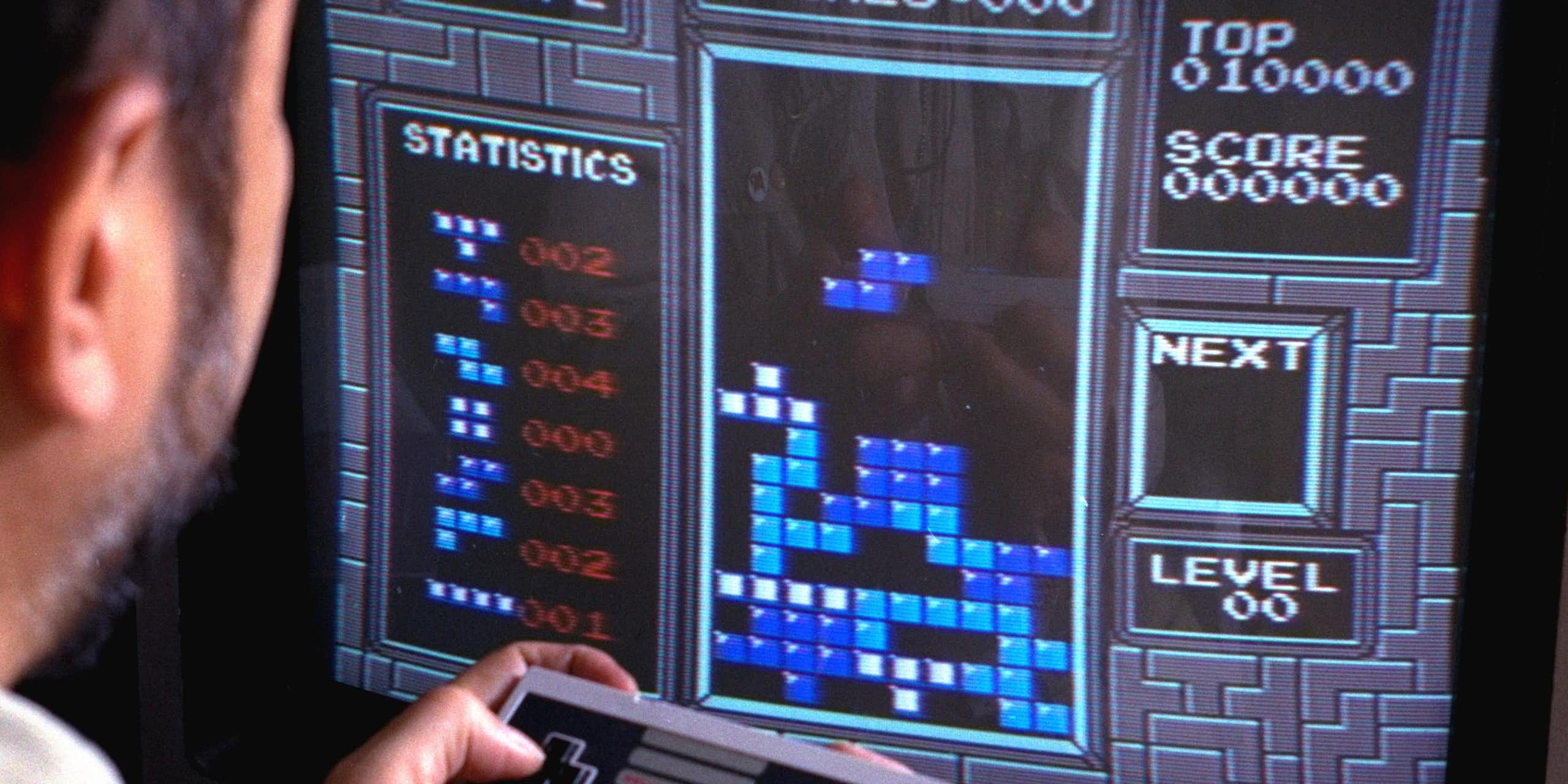 A man sitting in front of a TV screen displaying a 'Tetris' board with shapes stacking on top of each other. The man is holding a video game controller. 