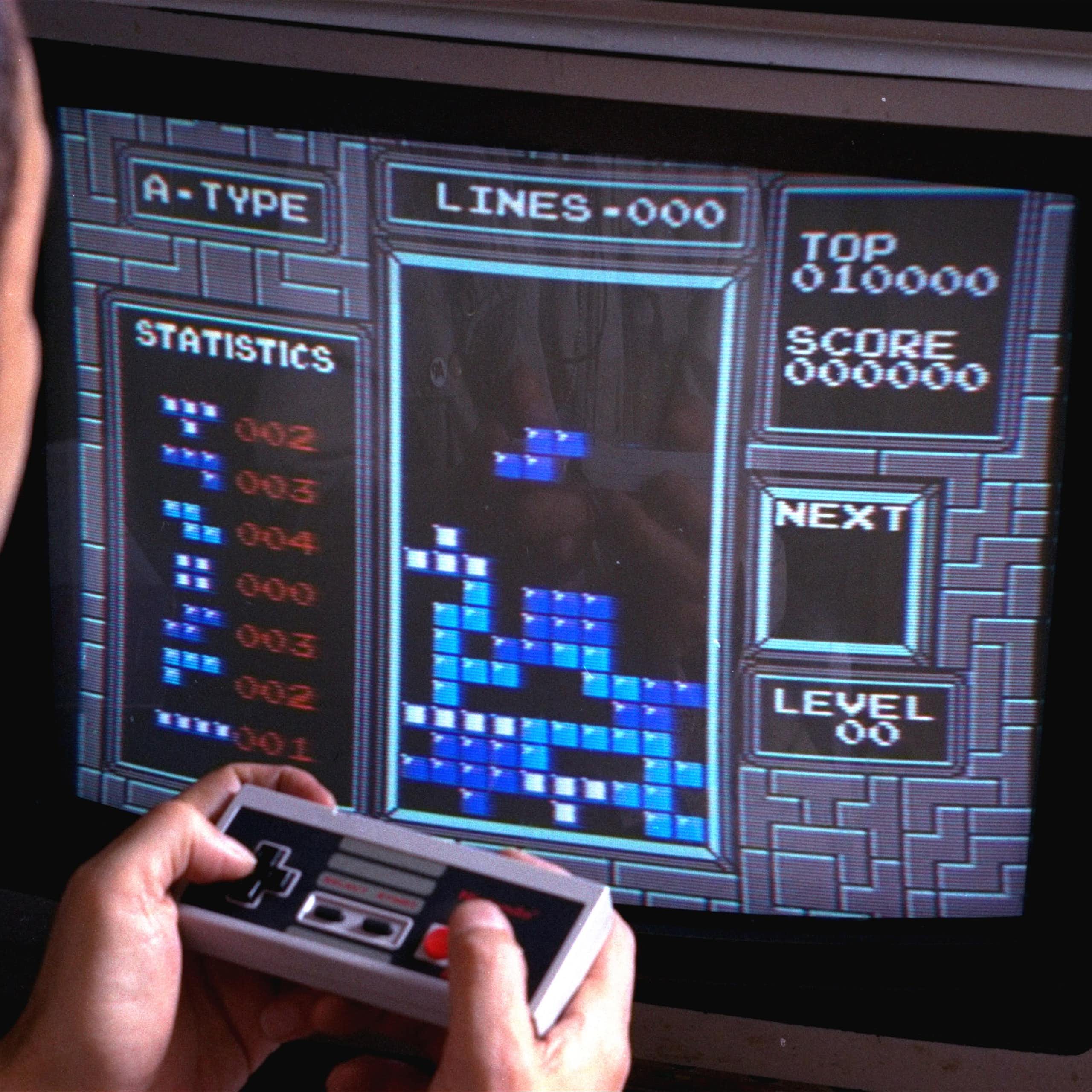 A man sitting in front of a TV screen displaying a 'Tetris' board with shapes stacking on top of each other. The man is holding a video game controller. 