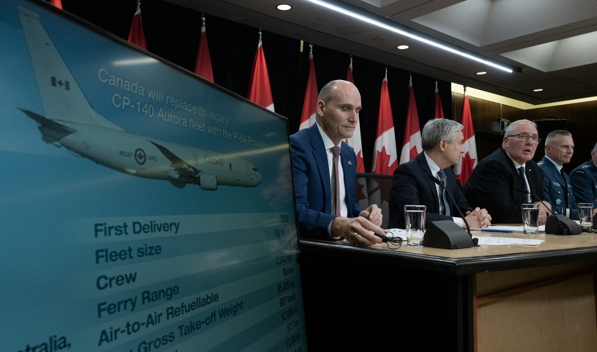 A row of men sit at a table next to a screen displaying surveillance planes.