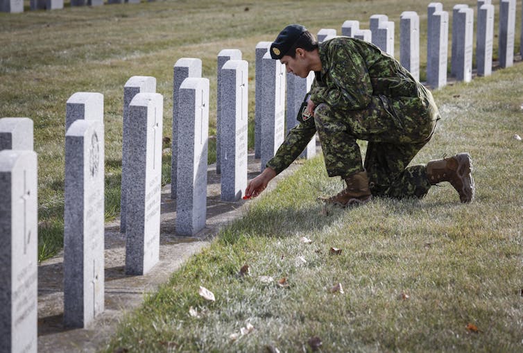 A man wearing camouflage kneels at a row of white gravestones and places a poppy at the foot of one of them.