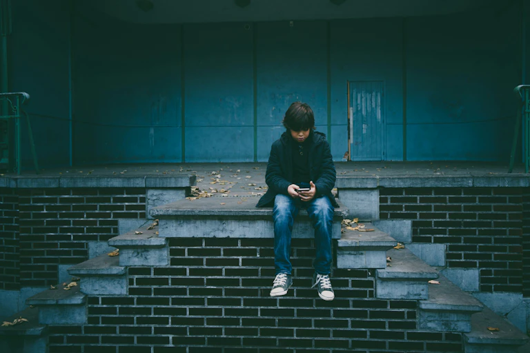 A boy sits on concrete stairs, dresses in jeans and sneakers, looking at a phone.