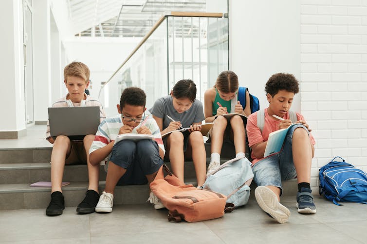 Five students sit on steps with backpacks, writing in books and working on a laptop.