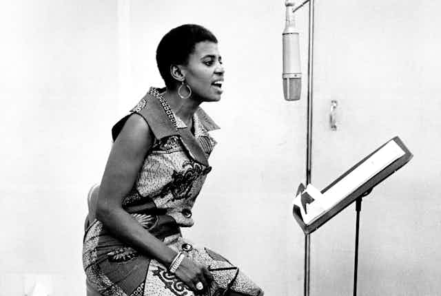 A black and white photo of a young African woman in a music studio in a sleeveless African print dress sitting and singing into a microphone in profile.