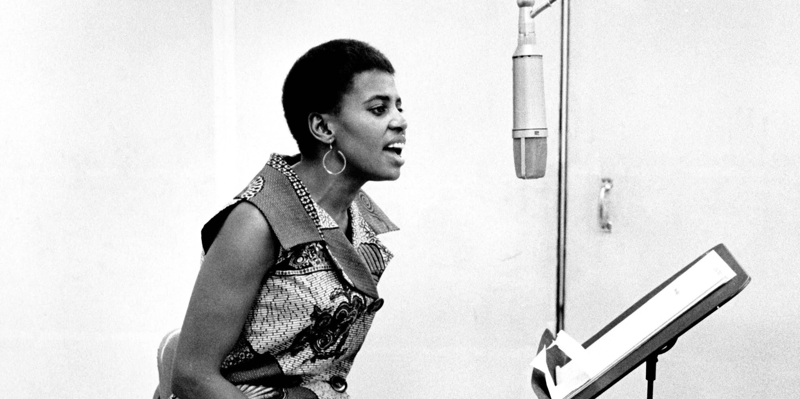 A black and white photo of a young African woman in a music studio in a sleeveless African print dress sitting and singing into a microphone in profile.