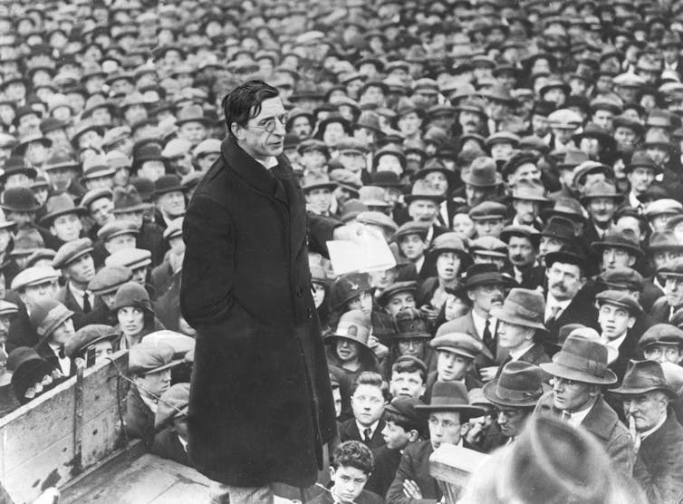Éamon de Valera standing on a stage with his back turned to a large crowd.