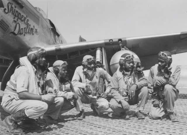 five black pilots crouching in front of a mustang plane