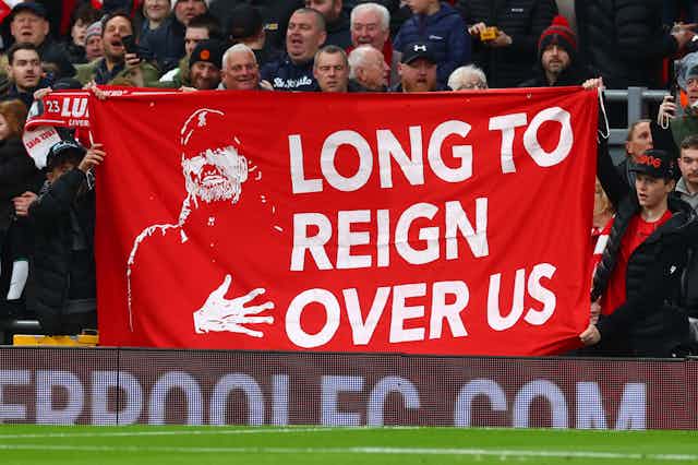 A group of Liverpool FC supporters hold up a large red and white banner depicting an image of manager Jurgen Klopp and the words 'Long to reign over us'