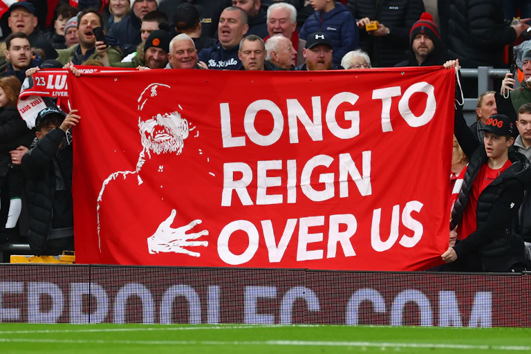 A group of Liverpool FC supporters hold up a large red and white banner depicting an image of manager Jurgen Klopp and the words 'Long to reign over us'
