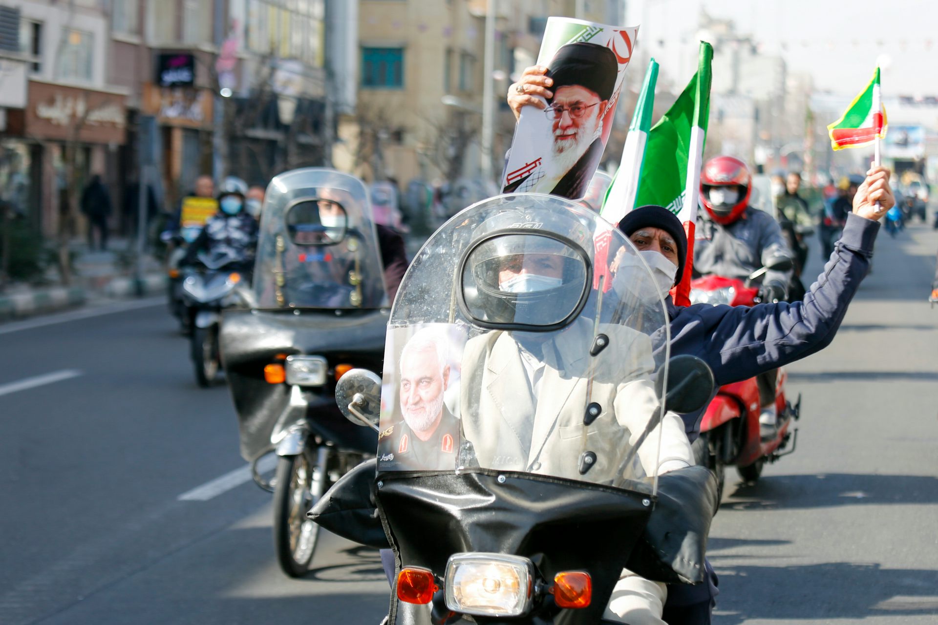 People driving their motorcycles down a city road holding Iranian flags and banners commemorating the Iranian Revolution.