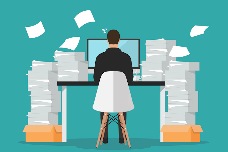 A person seen from the back, sitting at a computer with piles of paper around them.
