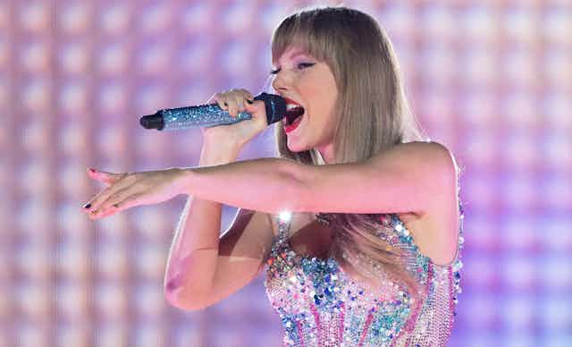 Taylor Swift singing into a microphone and pointing onstage in her Eras tour, wearing a sequined top