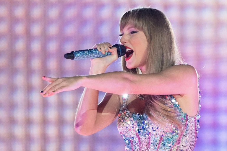 Taylor Swift singing into a microphone and pointing onstage in her Eras tour, wearing a sequined top