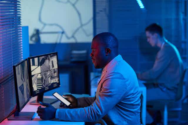 Two men sit in front of computers and other screens in a darkened room