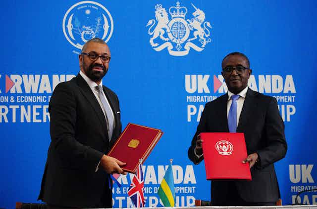 James Cleverly and Vincent Biruta holding red folders in front of a blue backdrop reading UK Rwanda partnership