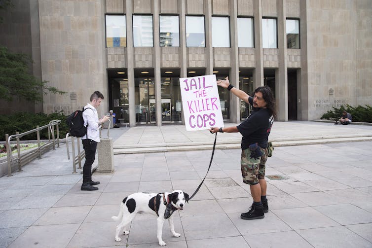 A man with a dog holds a sign that reads Jail the Killer Cops outside a court building.