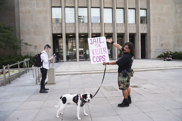 A man with a dog holds a sign that reads Jail the Killer Cops outside a court building.
