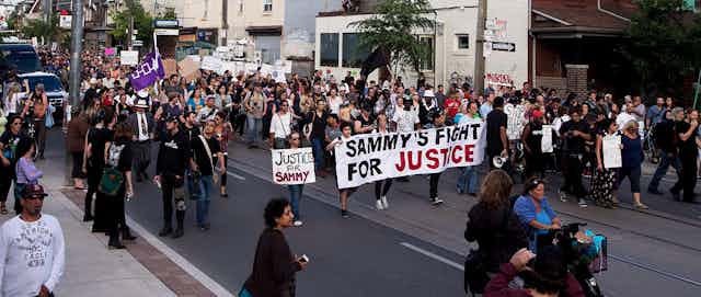 A crowd marches along a street carrying a banner that reads Sammy's Fight for Justice.