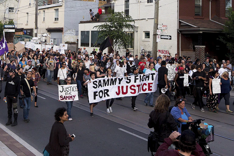 A crowd marches along a street carrying a banner that reads Sammy's Fight for Justice.