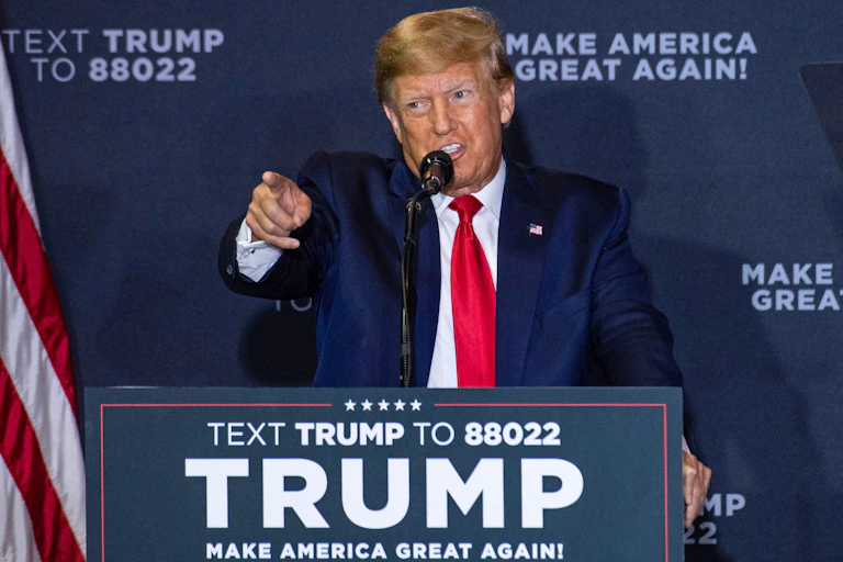 A sandy-haired man in a blue jacket, white shirt and red tie at a lectern that says 'TRUMP' on it.