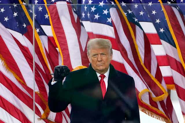 A man in an overcoat stands in front of American flags holding up a closed fist.
