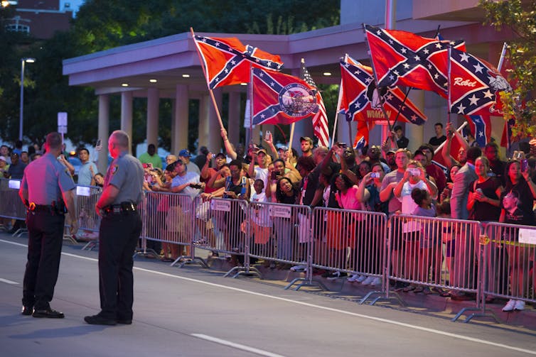 A crowd of people behind a barricade along a street, waving Confederate flags while watched by two police officers.