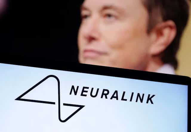 The blurry image of a man's face is seen behind the corner of a laptop screen displaying the word 'Neuralink'