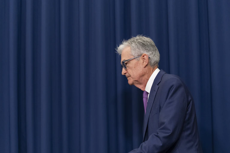 A man in glasses and suit walks in front of a blue curtain.