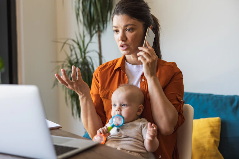 A young woman talks on a cell phone while looking at an open laptop. A baby sits on her lap chewing on a toy.