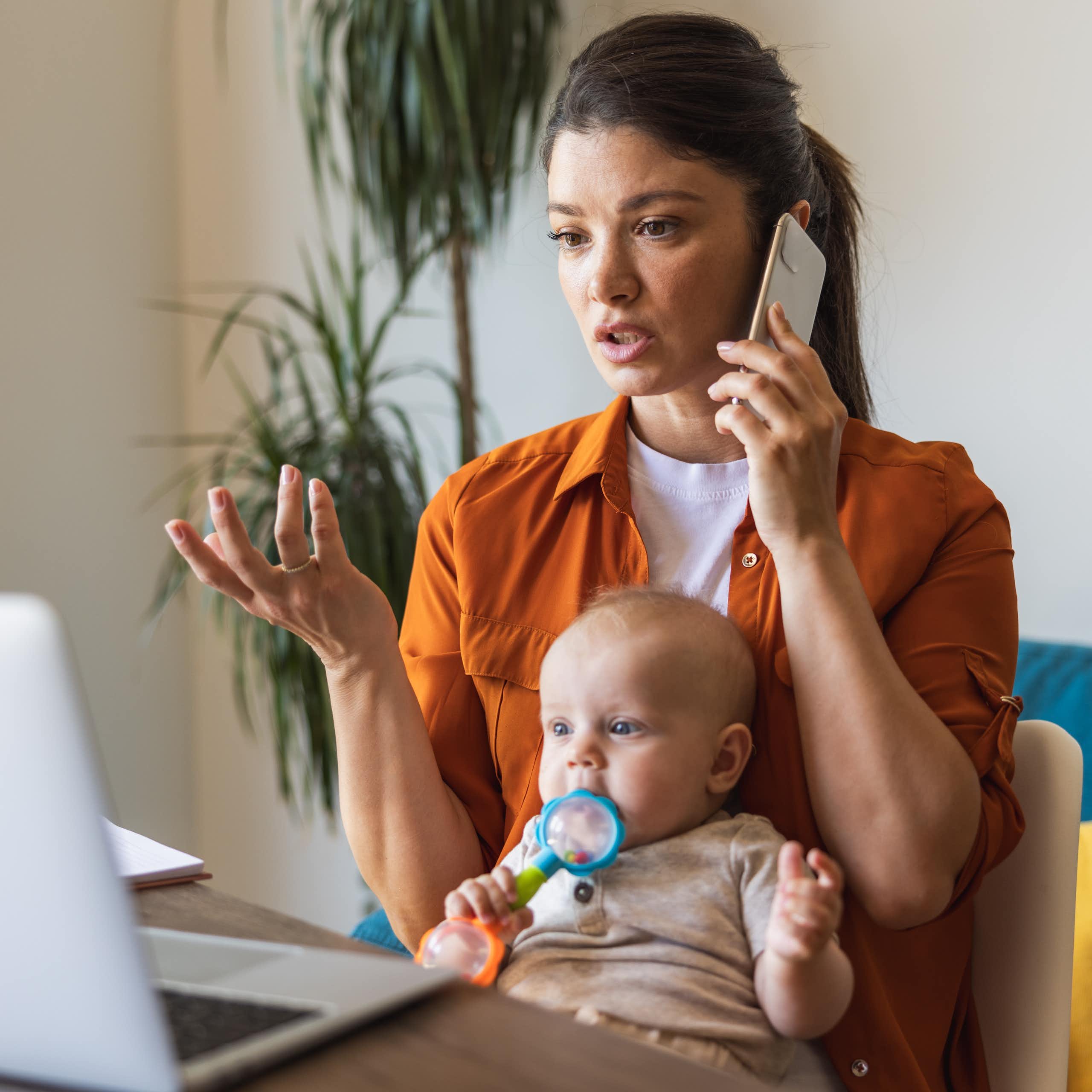 A young woman talks on a cell phone while looking at an open laptop. A baby sits on her lap chewing on a toy.
