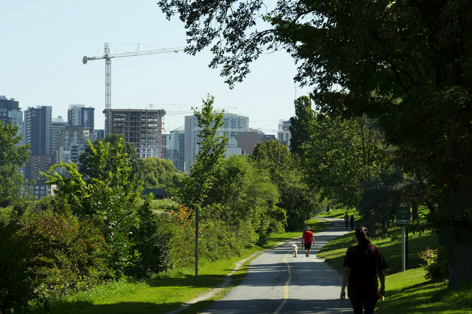 People walk down a path lined with greenery and trees while a city skyline looms in the background. A construction crane is seen beside a condo building that is under construction.