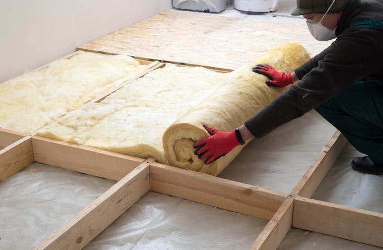 A worker in overalls unrolling insulation foam in the eaves of a house.