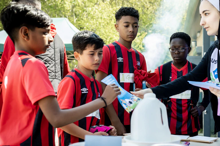 Young amateur football players in red and black kit on left, taking leaflets from two older students on the left side from across the table