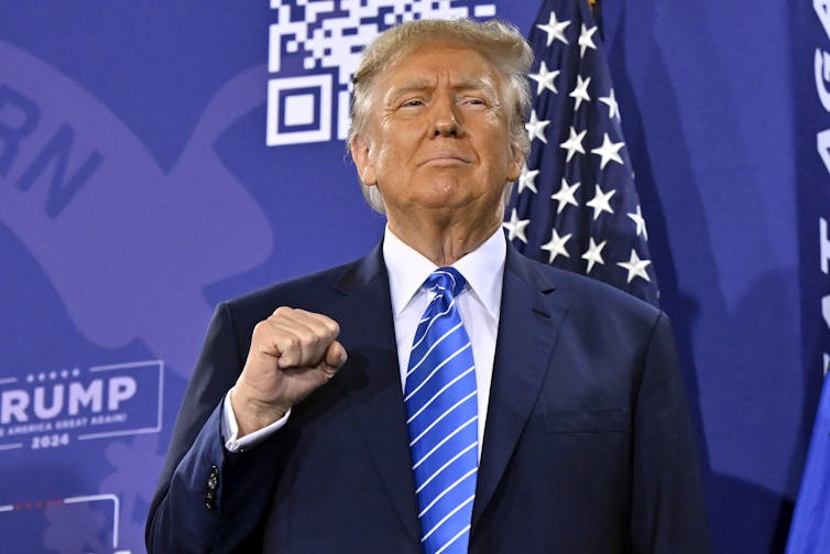 A man in a blue blazer, blue tie and white shirt in front of an American flag, holding his right hand in a fist.
