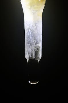 A photo of a crystalline tube with a drop of water at the end, against a black background.