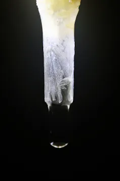 A photo of a crystalline tube with a drop of water at the end, against a black background.