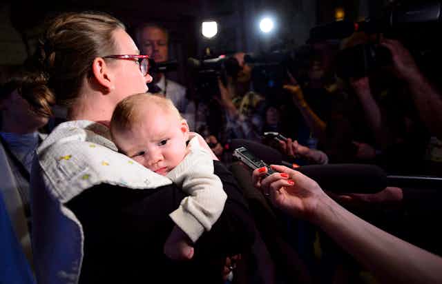 A woman wearning glasses holds a baby while in the glare of media lights.