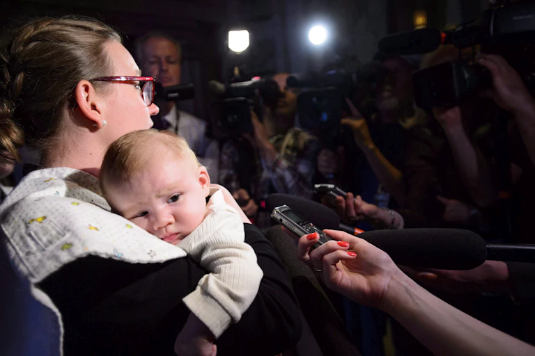 A woman wearning glasses holds a baby while in the glare of media lights.