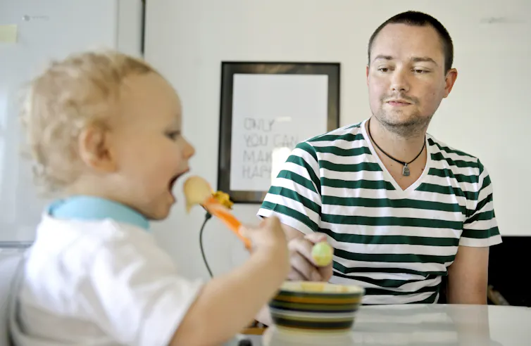 A man in a striped T-shirt watches his son eat in his high chair.