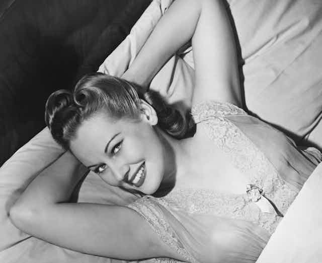 Black and white image, taken from an elevated angle, of a smiling woman looking at the camera -- with her hair in fifties style and wearing lace trimmed nightwear -- is in bed with both arms behind her head. The bedclothes are pulled up to her chest.