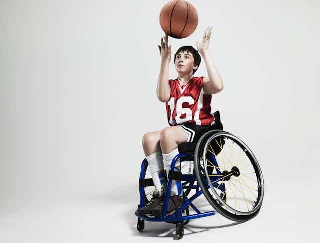 A boy in a wheelchair spins a basketball on his finger. He's wearing a red jersey with the number 16 in white numbers. And dark shorts.