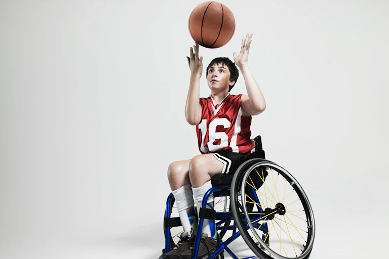 A boy in a wheelchair spins a basketball on his finger. He's wearing a red jersey with the number 16 in white numbers. And dark shorts.