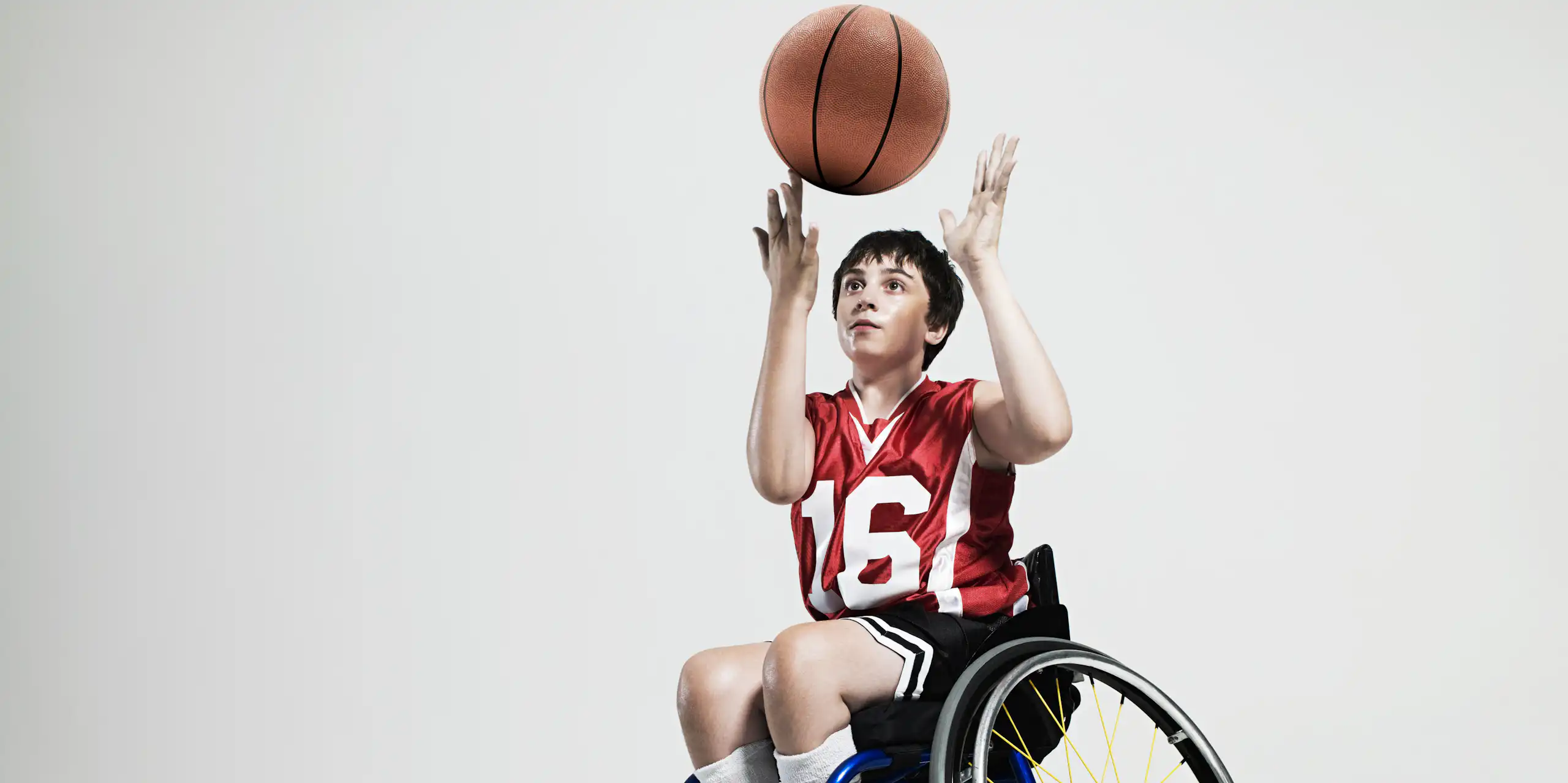 A boy in a wheelchair spins a basketball on his finger. He's wearing a red jersey with the number 16 in white numbers. And dark shorts.