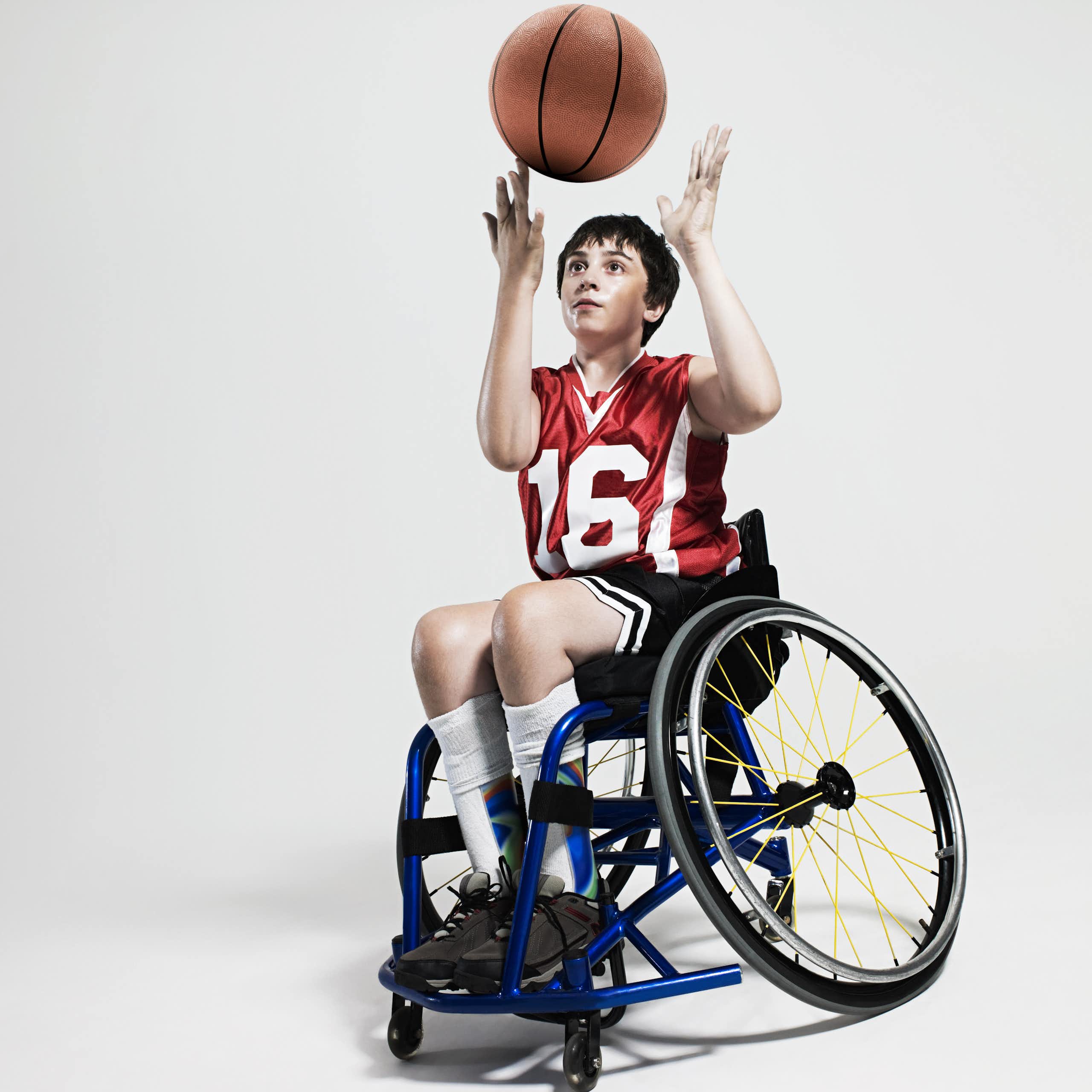 A boy in a wheelchair spins a basketball on his finger. He's wearing a red jersey with the number 16 in white numbers. And dark shorts.