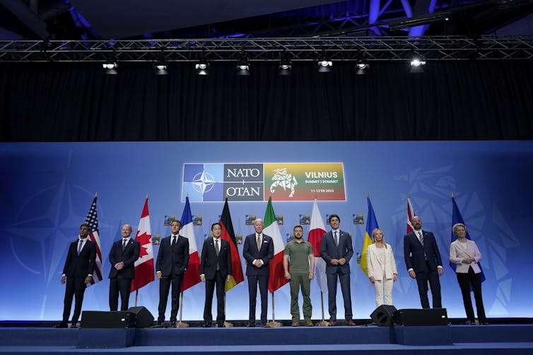A group of people stand in front of flags against a blue background.