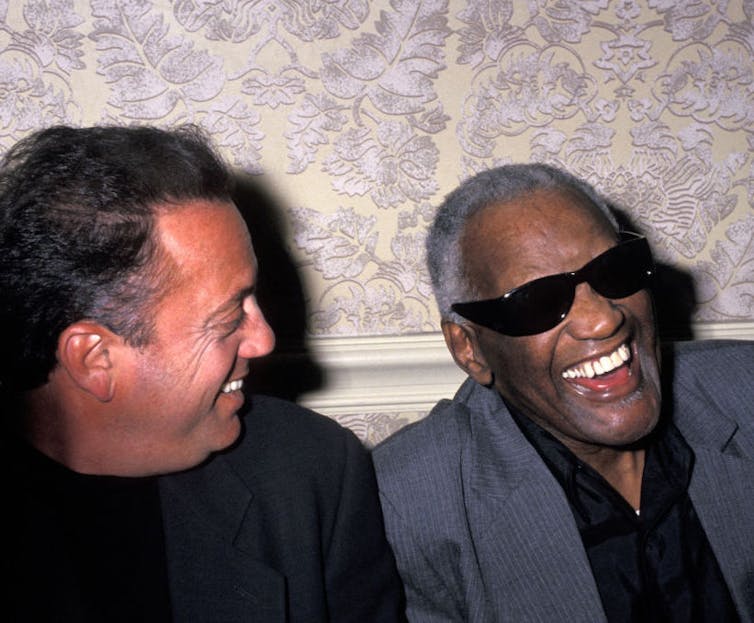 A man smiles and faces another man wearing black glasses who's howling with laughter.