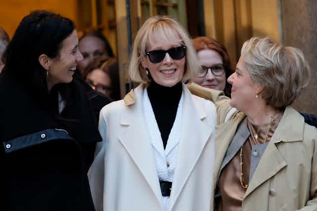 A woman in a white jacket with black sunglasses smiles, and walked arm in arm with two other women dressed in formal clothing.