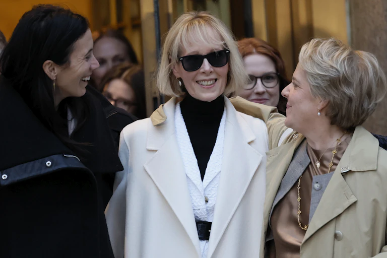 A woman in a white jacket with black sunglasses smiles, and walked arm in arm with two other women dressed in formal clothing.
