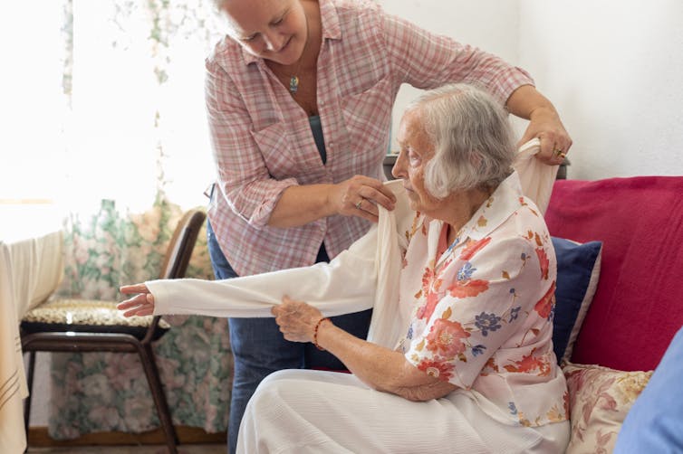 Mature woman places a cardigan on an elderly adult.