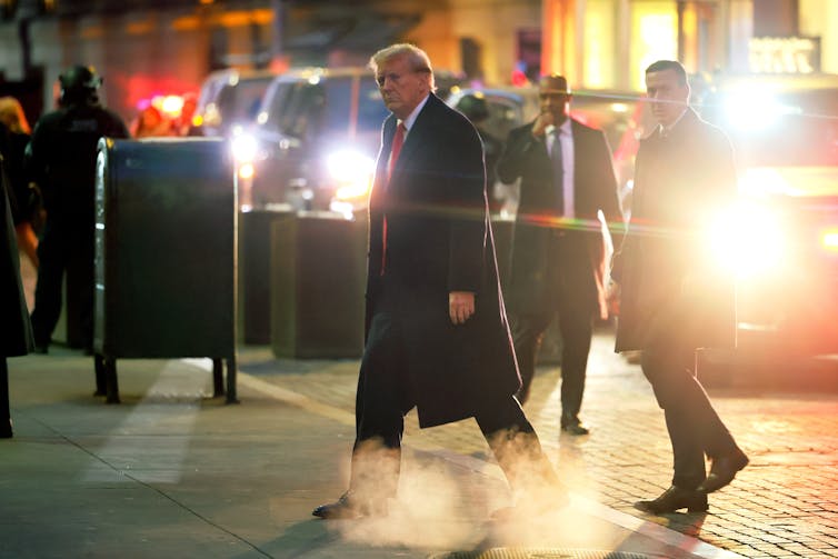 Donald Trump wears a long, black jacket and red tie and walks in a New York City street.