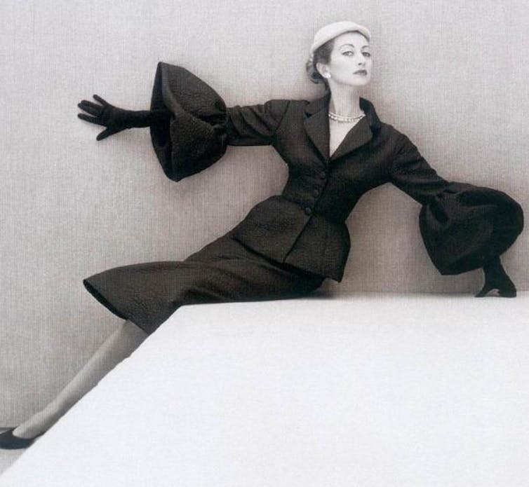 Woman wearing a black suit with flared bell sleeves and knee-length skirt sitting on a plinth with her right hand raised and pressed against the wall